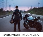 A biker stands next to a motorcycle on an asphalt road and looks into the distance. Summer sunset