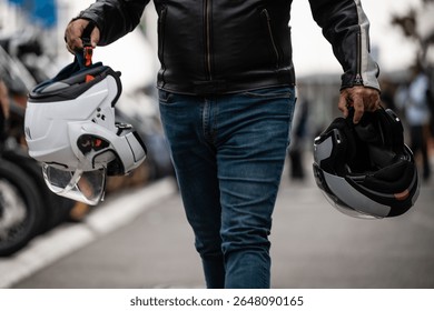 Biker walking while carrying two helmets at a motorcycle event, wearing a leather jacket and jeans
