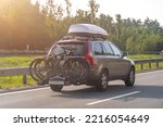 Bikes fastened on bicycle holder mounted on back side of car on country road. Brown car with roof luggage box and trunk bike rack driving on highway.