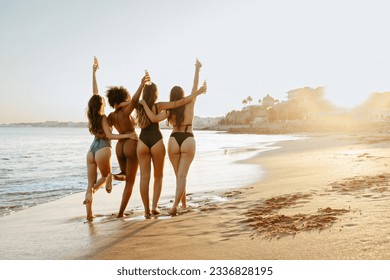 Bikini fun. Group of diverse women in swimwear embracing and walking at the beach, enjoying sunset, copy space, back view. Carefree female friends making happy memories during summer vacation - Powered by Shutterstock