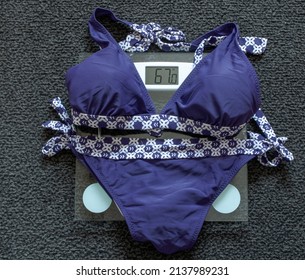 Bikini On A Glass Scale Which Marks 67 Kg, Dark Gray Carpet In Background