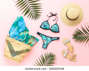 Bikini swimsuit with tropical print, silver glitter flat sandans, straw hat, wicker beach bag, sarong, tropical palm leaves on pink background. Overhead view of woman's swimwear and beach accessories.