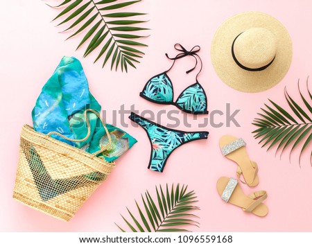 Bikini swimsuit with tropical print, silver glitter flat sandans, straw hat, wicker beach bag, sarong, tropical palm leaves on pink background. Overhead view of woman's swimwear and beach accessories.