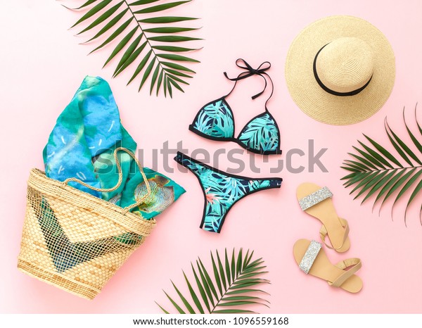 Bikini swimsuit with tropical print, silver\
glitter flat sandans, straw hat, wicker beach bag, sarong, tropical\
palm leaves on pink background. Overhead view of woman\'s swimwear\
and beach accessories.