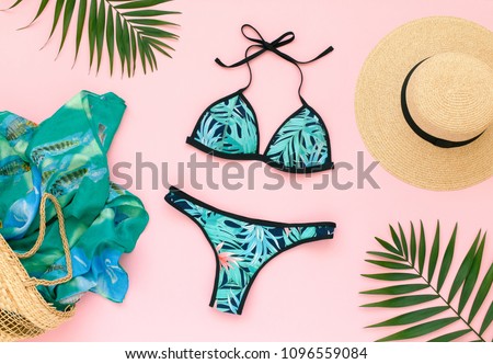 Bikini swimsuit with tropical print, straw hat, wicker beach bag, sarong and tropical date palm leaves on pink background. Overhead view of woman's swimwear and beach accessories. Flat lay, top view.