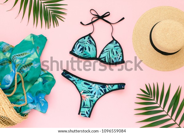 Bikini swimsuit with tropical print, straw hat,\
wicker beach bag, sarong and tropical date palm leaves on pink\
background. Overhead view of woman\'s swimwear and beach\
accessories. Flat lay, top\
view.
