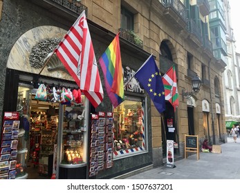 Bilbao Spain. Four Large Flags Overhanging A Shop Window. Atletico Bilbao Football Club. Gay Pride. European Union. Basque National Flag. In Narrow Cobbled Alleyway Of The Old Quarter