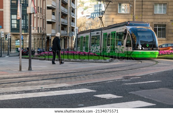 BILBAO, SPAIN-DECEMBER 19, 2021 : Elderly people\
walking near Euskotren city tram runs on tramway track. Modern\
transport in Europe. Sidewalk in smart city. Electric vehicle.\
Sustainable travel.