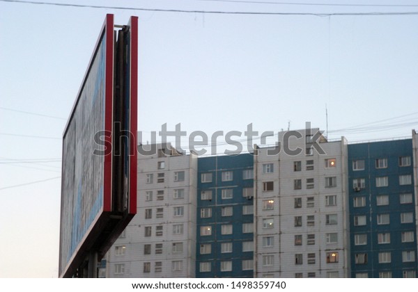 Billboard of bright red color a look with a side\
on the city street against the background of the blue and White\
multi-storey building with square windows and the gray sky,\
electric wires.