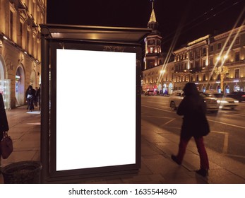 Billboard In A Bus Stop. Glowing Box With An Advertising Poster Standing In The City At Night. With People Walking And Riding Cars