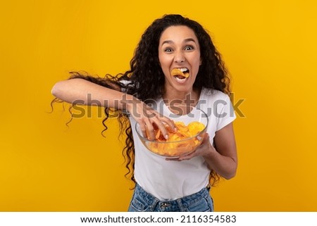 Binge Eating Concept. Portrait Of Funny Hungry Woman Enjoying Delicious Potato Crisps Holding Glass Bowl, Posing With Chips In Mouth Isolated Over Yellow Orange Studio Wall. Junk Meal Addiction