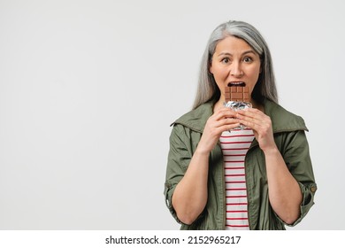 Binge Eating, Overeating. Unhealthy Junk Food. Calories And Saturated Fats. Mature Middle-aged Woman With Grey Hair Eating Chocolate Bar, Hungry During PMS Menopause Climax Isolated In White