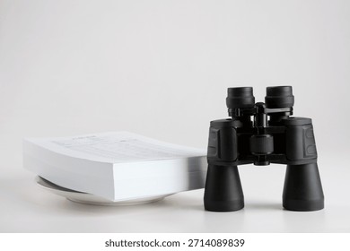 Binoculars placed next to a stack of papers on a flat surface in a simple setting during indoor daylight hours