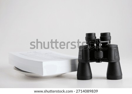 Binoculars placed next to a stack of papers on a flat surface in a simple setting during indoor daylight hours