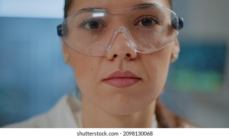 Biochemist With Safety Goggles And White Coat In Laboratory, Face Of Scientist Looking At Camera While She Works On Chemistry Development In Lab. Portrait Of Woman Doing Research. Close Up