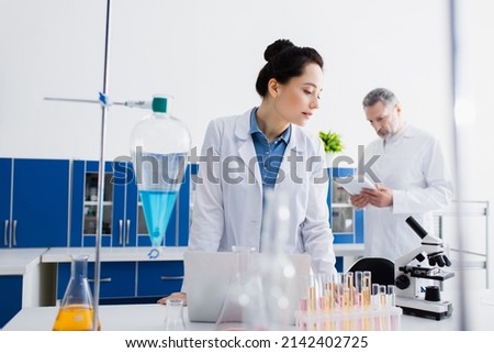 bioengineer looking at microscope while standing near test tubes and flasks in lab