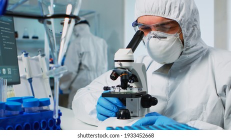 Biological scientist in coverall using microscope doing research for new vaccine in pharmaceutical labolatory. Coworkers examining virus evolution using high tech researching diagnosis against covid19