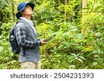 A biologist looks up at the trees above him, spotting species needed for his research. He holds a tablet with data, connected to the internet for troubleshooting, in the jungle