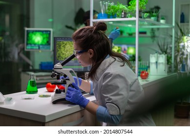 Biologist scientist in white coat working in expertise laboratory looking into microscope analyzing organic gmo leaf. Specialist researcher doing biochemistry experiment.