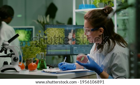 Biologist woman writing medical expertise while holding petri dish with vegan beef meat in hands working in microbiology laboratory. Chemist woman researching food modified genetically