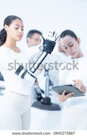 Bionic engineering workshop with eyes down, the note taker working through her tablet entries while the raised prosthetic arm stays lit against the studio wall.