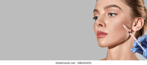 Biorevitalization procedure. Pretty woman while beauty injections with hyaluronic acid for smoothing of mimic wrinkles around eyes, over grey background with space for text - Shutterstock ID 2081453251