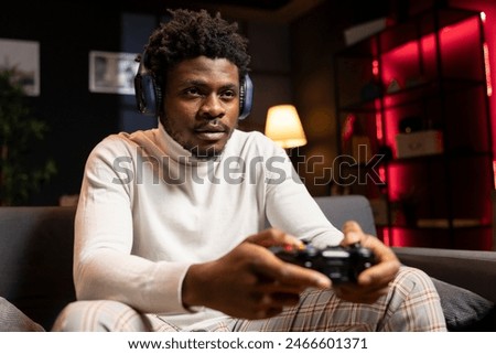 BIPOC man playing videogames using controller, having fun in red neon lit apartment. Gamer competing in online multiplayer game on gaming console, using headphones to talk with teammates