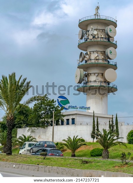 Bir Morad Raîs, Algeria - March 11, 2022: Algérie\
Télécom telecommunication antenna tower with the brandmark logo of\
the phone and internet provider national company. Cars, grass and\
palm trees. 