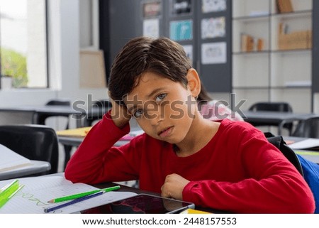 Biracial boy with brown hair looks sad in a school classroom setting. Dressed in a red shirt, he rests his head on his hand, surrounded by school supplies.