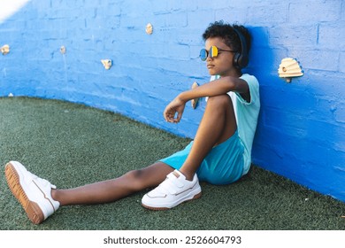 Biracial boy enjoys music outdoors, with copy space. He relaxing against a blue wall while wearing headphones and sunglasses. - Powered by Shutterstock