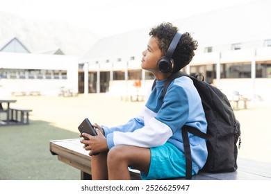 Biracial boy enjoys music at school, with copy space. He seated outdoors with headphones, engrossed in his smartphone. - Powered by Shutterstock