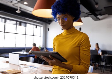 Biracial casual businesswoman with blue afro using tablet in casual office. Casual office, business, communication and creative work, unaltered. - Powered by Shutterstock