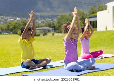 Biracial children practice yoga outdoors, sitting on colorful mats. The 9-year-old girl in purple leads the 8-year-old boy in yellow and the 10-year-old girl in pink in a peaceful pose. - Powered by Shutterstock