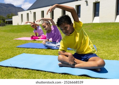 Biracial children practice yoga outdoors, with a boy in yellow leading the stretch. They are focused on their poses, sitting on colorful mats against a backdrop of greenery and buildings. - Powered by Shutterstock