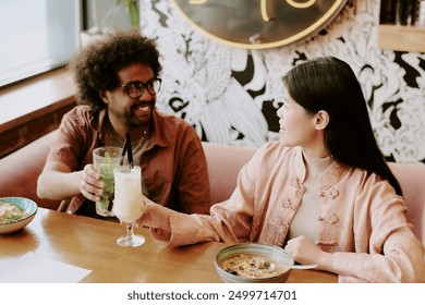 Biracial couple enjoying delicious Asian cuisine while having drinks at restaurant, smiling and talking joyfully. Both are seated at wooden table, natural light from window illuminating scene - Powered by Shutterstock