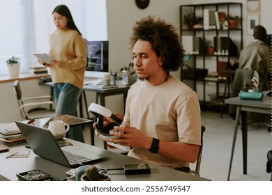 Biracial curly haired highly skilled IT specialist sitting at work desk holding VR headset and looking at laptop screen thoughtfully - Powered by Shutterstock