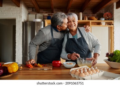 Biracial elderly couple happily cooking in the kitchen. Husband kissing wife on the cheek, healthy retired lifestyle. - Powered by Shutterstock