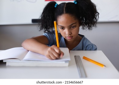 Biracial elementary schoolgirl writing on book at desk while studying in classroom. unaltered, education, learning, studying, concentration and school concept. - Powered by Shutterstock