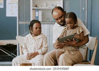 Biracial father spending quality time with daughters teaching them using tablet in living room creating a bonding moment while sitting together on chairs - Powered by Shutterstock