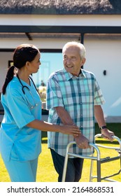 Biracial Female Doctor Helping Caucasian Senior Man In Walking With Walker In Yard On Sunny Day. House, Assistance, Unaltered, Healthcare, Patient, Retirement, Support, Recovery, Disability Concept.