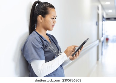 Biracial female doctor wearing scrubs using tablet in hospital corridor. Medicine, healthcare, communication, work and hospital, unaltered. - Powered by Shutterstock