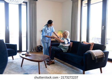 Biracial Female Health Worker Helping Caucasian Senior Man To Get Up From The Couch At Home. Medical Care And Retirement Senior Lifestyle Concept