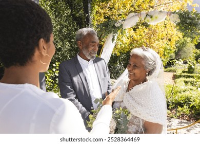 Biracial female marriage officiant and happy senior couple during wedding in sunny garden. Love, ceremony, marriage, summer and celebration, unaltered. - Powered by Shutterstock