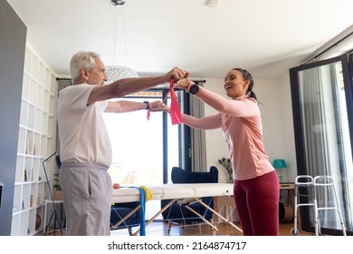 Biracial female physiotherapist with caucasian senior man stretching resistance band at home. Assisting, unaltered, physical therapy, exercise, strength, healthcare, retirement, training, recovery. - Powered by Shutterstock