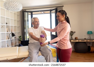 Biracial female physiotherapist helping caucasian senior man lifting dumbbell while sitting at home. Assistance, unaltered, physical therapy, exercise, healthcare, retirement, treatment, recovery. - Powered by Shutterstock