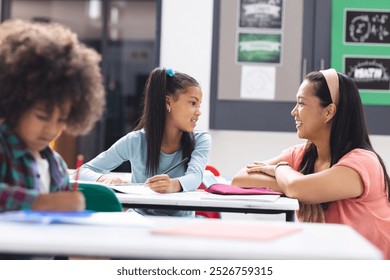 Biracial female teacher interacting with biracial female student in classroom. Another student studying at desk in bright, modern school environment, unaltered - Powered by Shutterstock