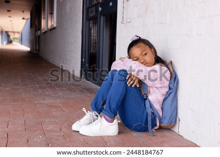 Biracial girl appears sad, sitting alone against a wall with copy space in school. Her downcast eyes and slumped posture suggest loneliness in an outdoor setting.