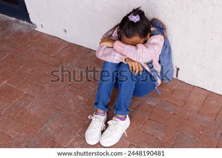 Biracial girl appears upset, sitting on the ground with her head down, with copy space. She is wearing a plaid shirt and jeans, conveying a sense of distress or sadness.