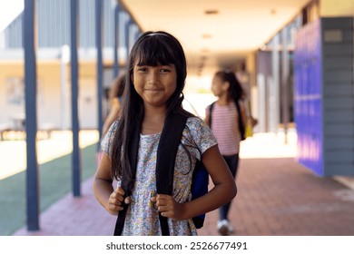 Biracial girl with long dark hair smiles in school, holding her backpack straps. She stands in a school corridor, with two peers chatting in the background. - Powered by Shutterstock