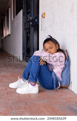 Biracial girl with a sad expression sits hugging her knees in school. She wears a pink and blue checkered shirt, denim overalls, and white sneakers, conveying a casual style.